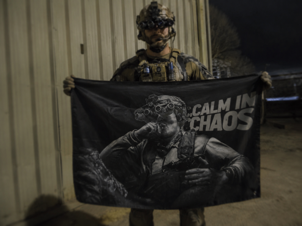 Person in military gear holding a flag with a graphic and text 'CALM IN CHAOS' in a dimly lit setting.